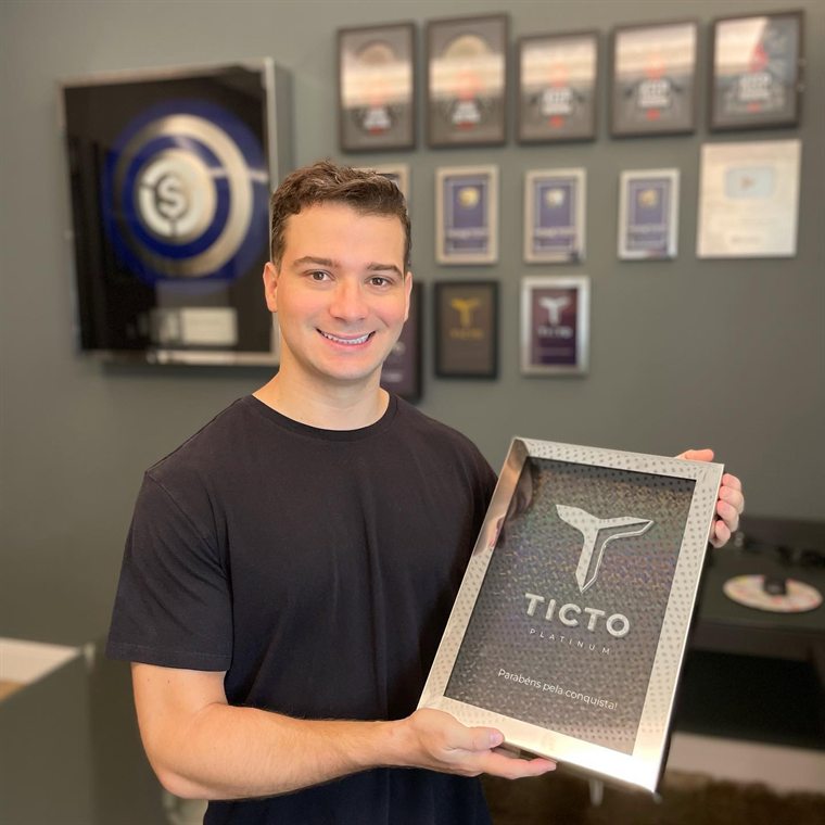 João Pedro segurando a placa Platinum da Ticto, com outras premiações na parede